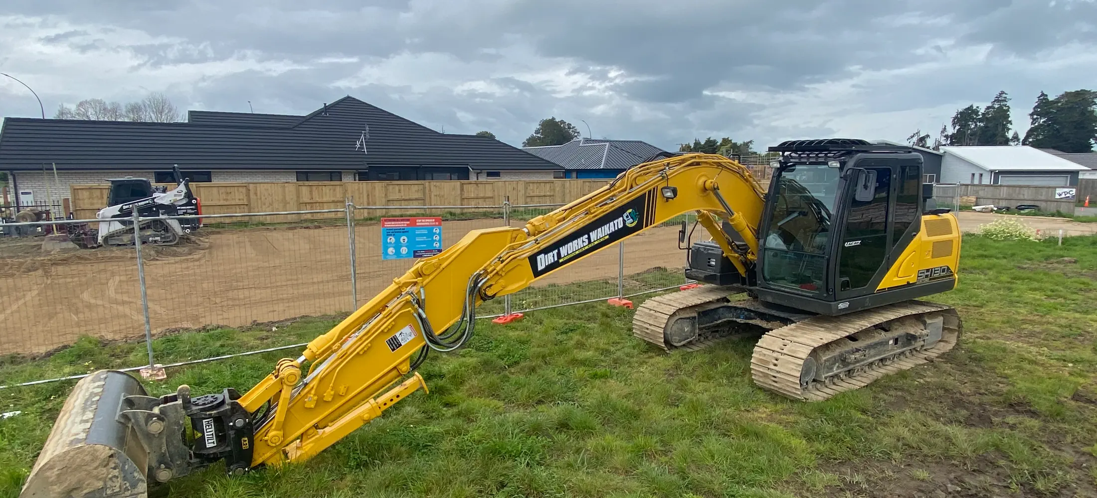 Excavator on a fenced residential lot with its boom laid out and the bucket off, houses behind