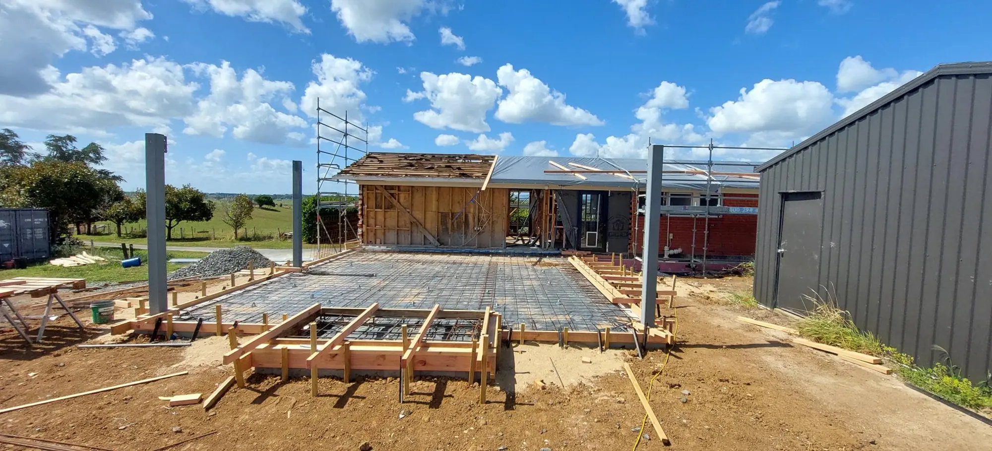 New house slab boxed and poured, with the timber-framed shell going up on it beside a farm shed