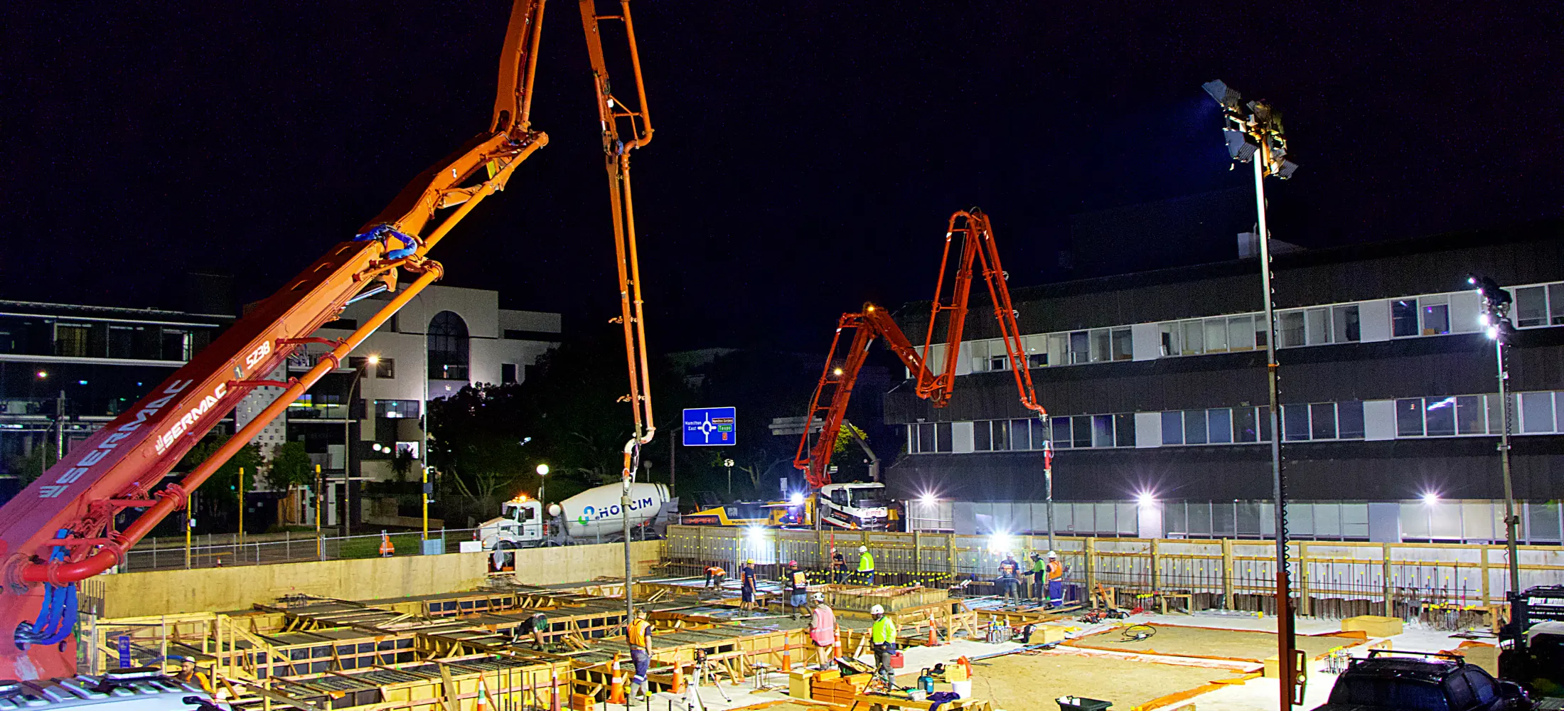 Two concrete pump booms lit up over a commercial pour running through the night in town
