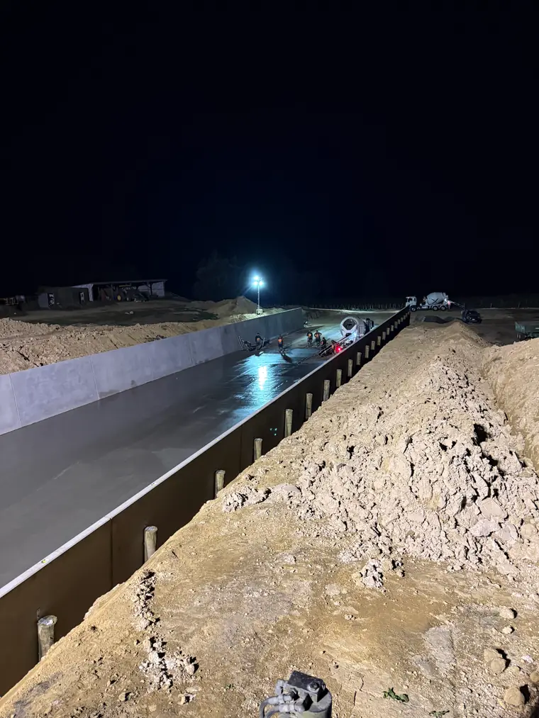 Long concrete channel poured beside the cut bank, still wet under floodlight
