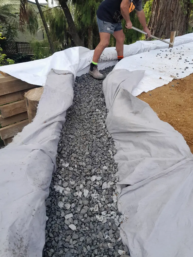 Worker raking drainage metal into a geotextile-lined subsoil drain beside a timber retaining wall