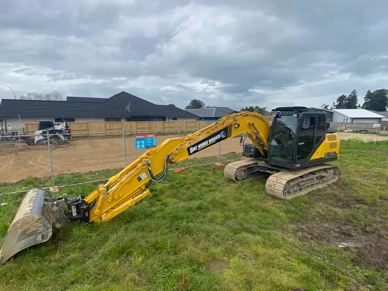 Excavator on a fenced residential lot with its boom laid out and the bucket off, houses behind